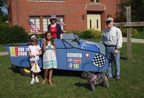 Five people stand in front of a decorated blue race car on a lawn beside a brick building.